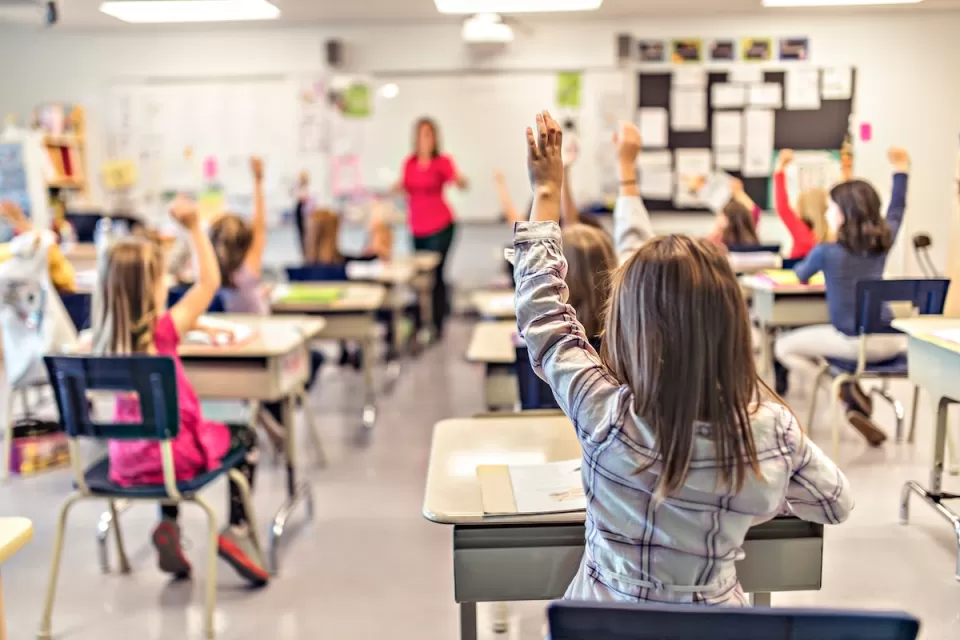 View from back of classroom of elementary school children at desks with raised hands engaged in class.